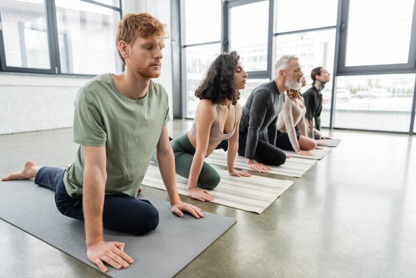 Kızıl saçlı adam yoga sınıfında çok ırklı insanların yanında Half Pigeon Asana egzersizi yapıyor.  - Fotoğraf, Görsel