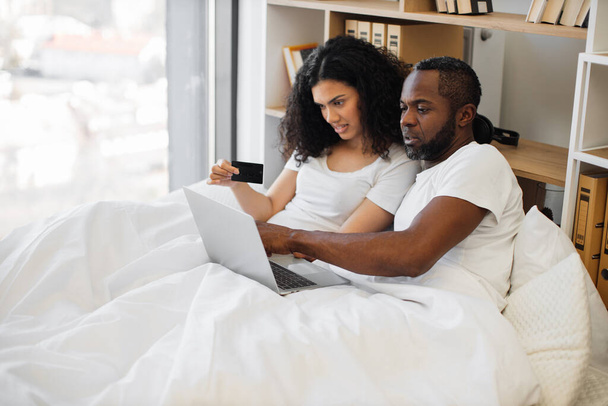 Loving multiethnic family spending morning in bed with laptop while using credit card for internet banking at home. Romantic partners making online payment operations in available application. - Foto, Imagen