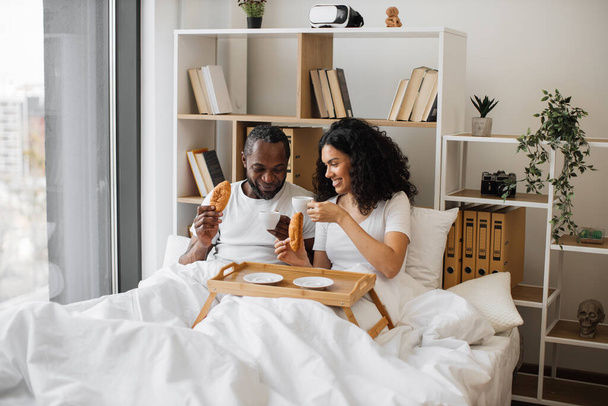 African american family man and woman eating crescent rolls and drinking black coffee while lying on bed in sunny morning. Joyful married couple sending message of true love through daily care. - Fotografie, Obrázek