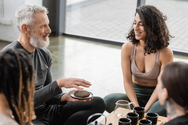 hombre tatuado con barba gris sosteniendo té puer comprimido cerca de alegre mujer de Oriente Medio en estudio de yoga  - Foto, imagen
