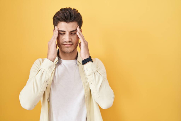 Jeune homme hispanique debout sur fond jaune avec la main sur la tête, maux de tête parce que le stress. migraine souffrant.  - Photo, image