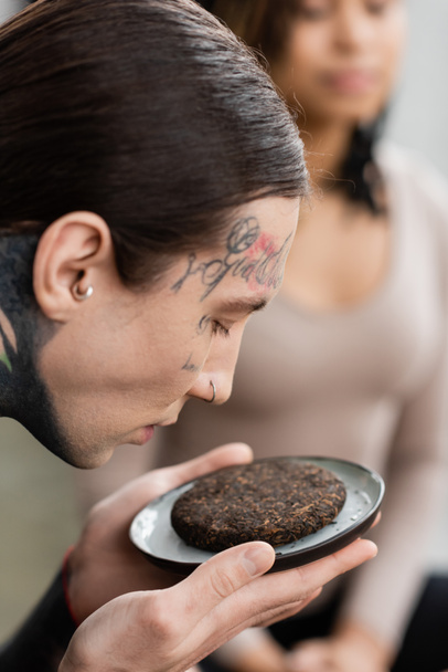 hombre joven y tatuado oliendo té puer fermentado cerca de una mujer afroamericana sobre un fondo borroso  - Foto, imagen