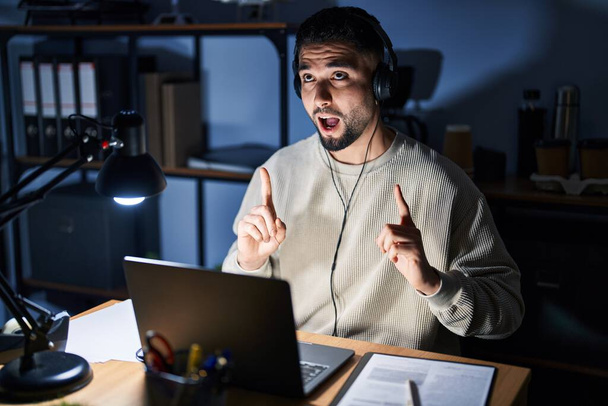 Młody przystojny mężczyzna pracujący w nocy za pomocą laptopa komputerowego zdumiony i zaskoczony patrząc w górę i wskazując palcami i podniesionymi ramionami.  - Zdjęcie, obraz
