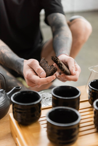 vista recortada del hombre tatuado rompiendo el té puer comprimido cerca de la olla de té tradicional china y tazas  - Foto, Imagen