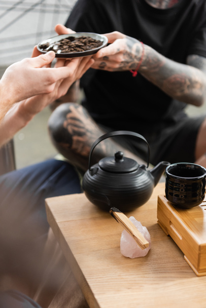 обрезанный вид на татуированного человека, проходящего компрессионный чайник рядом с традиционным чайником и чашками в студии йоги  - Фото, изображение
