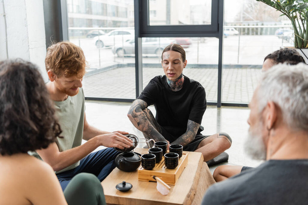 young man with tattoos sitting near interracial friends during tea ceremony in yoga studio  - Photo, Image