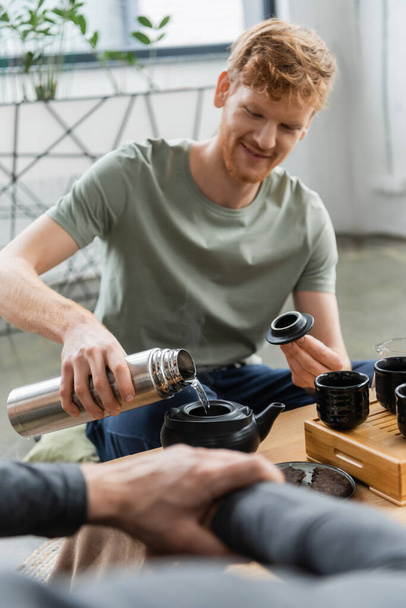 happy redhead man holding thermos while pouring hot water and brewing puer tea in Chinese tea pot  - Photo, Image