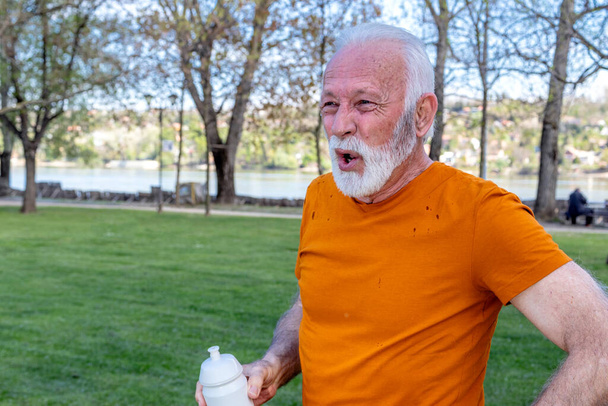 Un hombre mayor exhausto está recuperando el aliento después de hacer ejercicio en el parque público de la ciudad junto al río. Refrescante con agua de una botella de plástico. - Foto, imagen