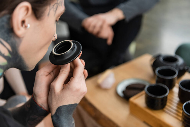 tattooed young man smelling puer tea near Chinese cups in yoga studio  - Photo, Image