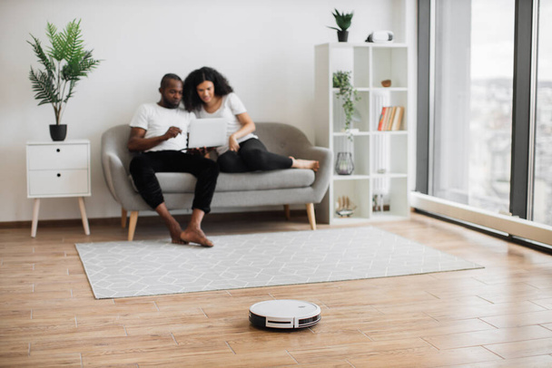 Automatic robot vacuum cleaning wooden floor while happy young couple resting on background. Two multicultural in love spending time for watching movie instead of households. - Foto, imagen