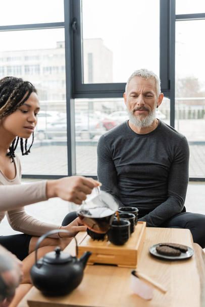 african american woman with dreadlocks pouring puer into traditional tea cups near middle aged man - Photo, Image