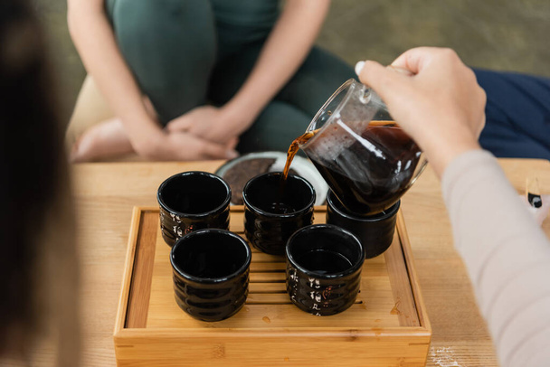 oříznutý pohled ženy nalévající puer ze skleněného džbánu do tradičních čajových šálků  - Fotografie, Obrázek