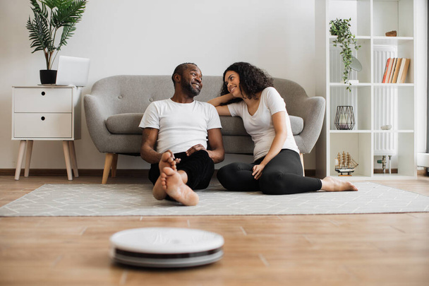 Relaxed multiethnic couple of two lovely people sitting on floor and using remote control for managing work of automated vacuum cleaner. Domestic chores with modern robot. - 写真・画像