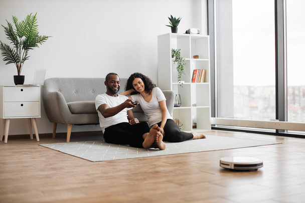 Relaxed multiethnic couple of two lovely people sitting on floor and using remote control for managing work of automated vacuum cleaner. Domestic chores with modern robot. - Foto, imagen
