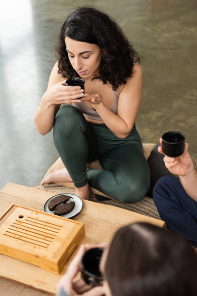 curly middle eastern woman smelling puer tea near men in yoga studio  - Foto, Bild