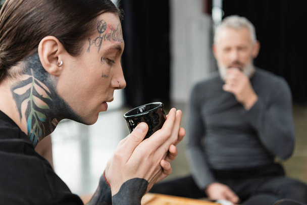 side view of tattooed man holding Chinese cup with brewed puer tea near middle aged man on blurred background  - Valokuva, kuva