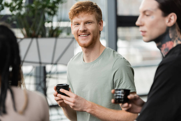 cheerful man with red hair holding japanese cup with puer tea near people in yoga studio  - Photo, image