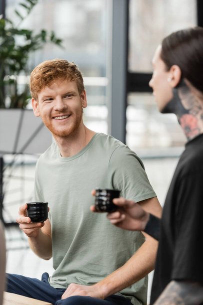 positive man with red hair holding Japanese cup with puer tea near tattooed friend in yoga studio  - Fotó, kép