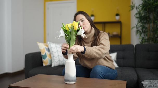 mooie latino vrouw zetten bloemen in een vaas zitten op de bank thuis - Foto, afbeelding