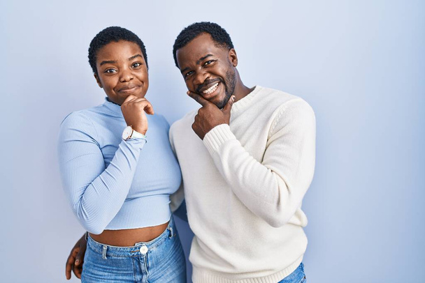 Fiatal afro-amerikai pár áll a kék háttér fölött, kezükkel az állukon, gondolkodva a kérdésről, elmélkedő kifejezésről. mosolygós és figyelmes arc. kétség.  - Fotó, kép