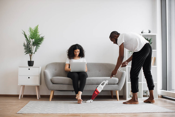 Side view of african man cleaning living room with cordless vacuum while focused woman working on laptop on couch. Caring husband doing households while wife freelancing at home. - Fotó, kép