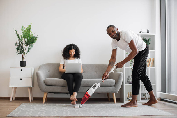 Side view of african man cleaning living room with cordless vacuum while focused woman working on laptop on couch. Caring husband doing households while wife freelancing at home. - Fotografie, Obrázek