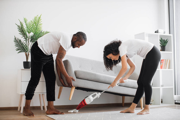Handsome caring man lifting grey couch for beloved wife for vacuuming underneath. Multicultural young couple in casual clothes cleaning modern apartment together during free time. - Zdjęcie, obraz