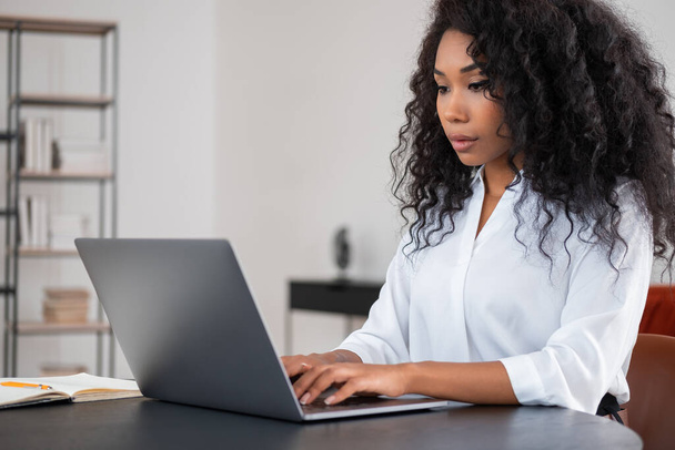 Black businesswoman typing on laptop, looking at the screen. Office room with shelf on blurred background. Concept of distance work and manager - Foto, Imagen