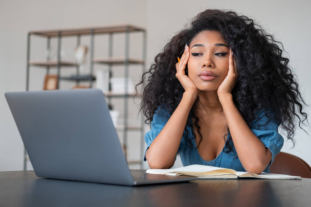 Stressed black businesswoman working with laptop, hands holding head and tired look. Office room with shelf on blurred background. Concept of question and answer - Фото, изображение