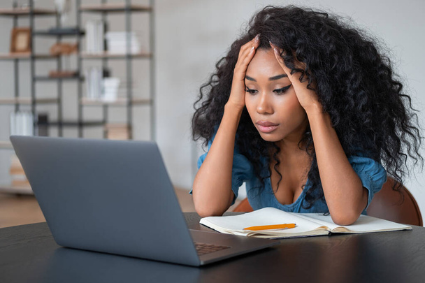 Stressed black businesswoman looking at laptop, hands holding head and upset look. Office room with shelf on blurred background. Concept of hardworking - Valokuva, kuva