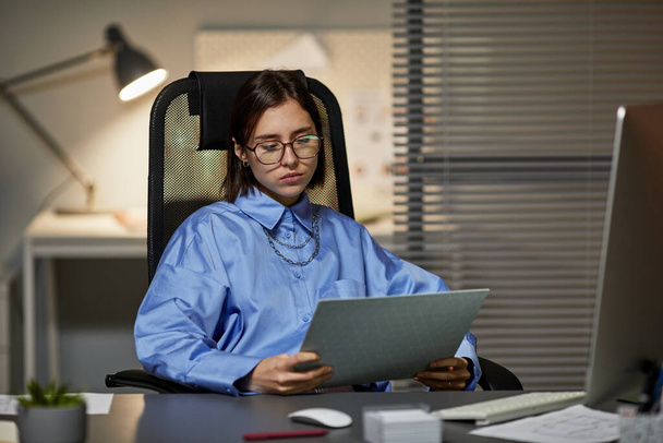 Ritratto di giovane imprenditrice seduta sul posto di lavoro e che legge documenti mentre lavora fino a tardi - Foto, immagini
