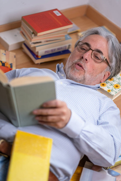 Hombre de pelo gris acostado cómodamente en el suelo lee fascinadamente un libro interesante y se sorprende de lo que está escrito - Foto, Imagen