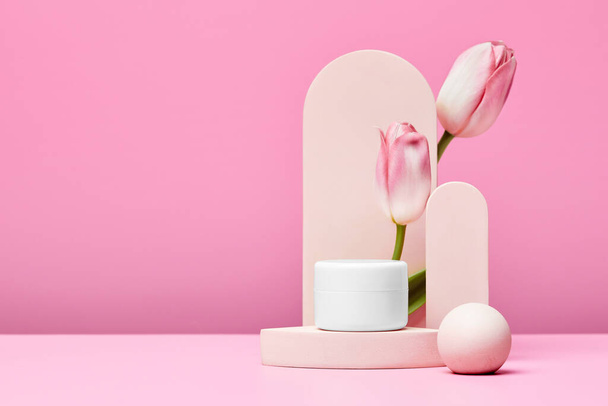 White jar of cosmetic premium product on podium with pink tulips and elegant arch on pastel pink background. mock up - Photo, Image