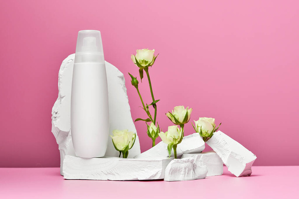 Bouteille blanche de crème ou lotion pour le corps avec pierres et roses blanches isolées sur fond rose - Photo, image