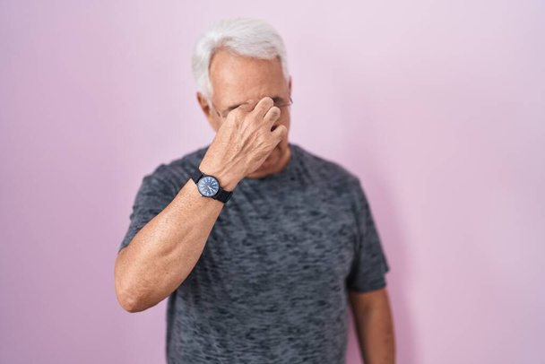 Homme d'âge moyen avec les cheveux gris debout sur fond rose fatigué se frottant le nez et les yeux ressentant fatigue et maux de tête. concept de stress et de frustration.  - Photo, image