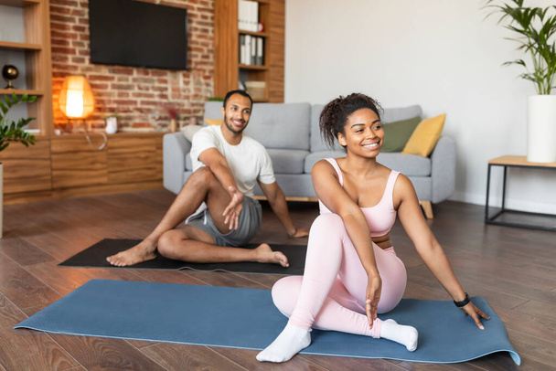 Молодые чернокожие муж и жена в одежде делают упражнения на растяжку, практикуют йогу, на коврике на полу в гостиной. Уход за телом и здоровьем, совместные тренировки, спорт и фитнес дома - Фото, изображение