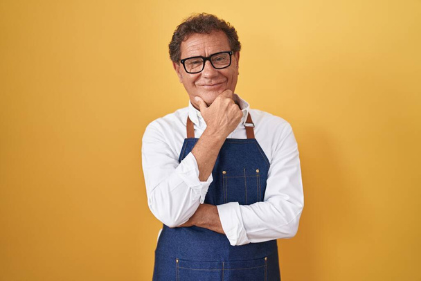 Middle age hispanic man wearing professional cook apron looking confident at the camera smiling with crossed arms and hand raised on chin. thinking positive.  - Valokuva, kuva