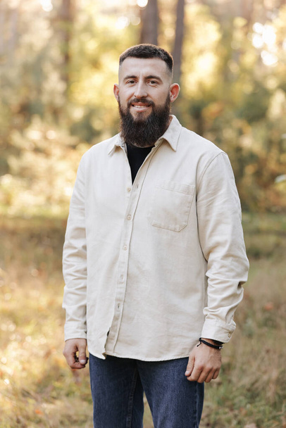 Retrato de jovem homem barbudo sorridente em jaqueta casual branco olha para a câmera no outono natureza fundo no campo ou no parque. .. Conceito de estilo, caminhando ao ar livre e unidade com a natureza - Foto, Imagem
