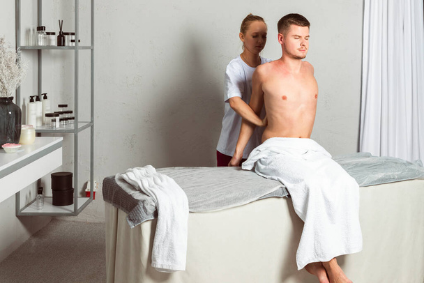 A professional masseuse or massage therapist is pulls the client's arms and shoulders, performing a stretch. the client lies covered with a towel on the massage table. - Photo, Image