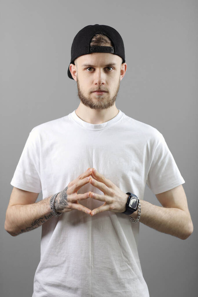 Stylish bearded caucasian young man with tattooed arm in cap, white t shirt and black pants is looking at camera, showing hand with ink picture on grey background. Fashion concept. - Foto, Imagem