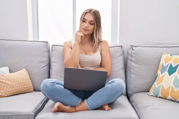 Blonde caucasian woman using laptop at home sitting on the sofa serious face thinking about question with hand on chin, thoughtful about confusing idea  - Photo, Image