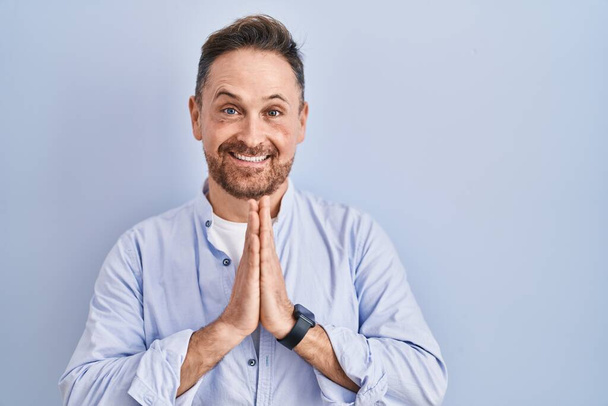 Middle age caucasian man standing over blue background praying with hands together asking for forgiveness smiling confident.  - Photo, Image
