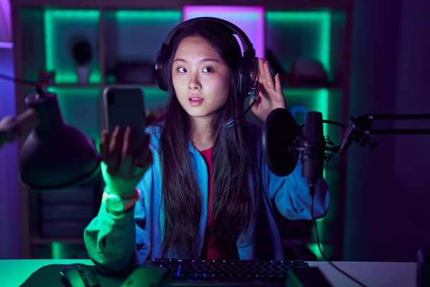 Giovane donna asiatica che gioca ai videogiochi con smartphone sorridendo con mano sull'orecchio ascoltando voci o pettegolezzi. concetto di sordità.  - Foto, immagini