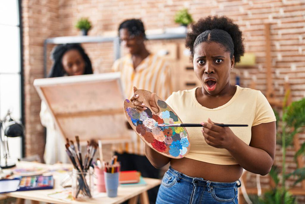 Joven pintora negra mujer en el estudio de arte sosteniendo paleta asustada y sorprendida con expresión sorpresa y asombro, miedo y cara excitada.  - Foto, imagen