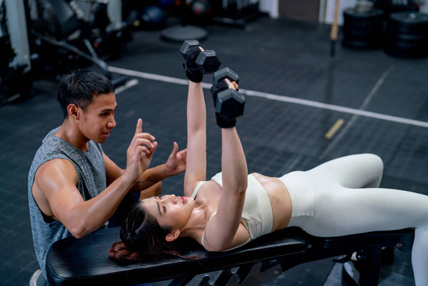 Asiatische Trainerin hilft und unterstützt asiatische Sportlerin beim Hantelheben mit Liegeposition auf langem Stuhl im Fitnessstudio. - Foto, Bild