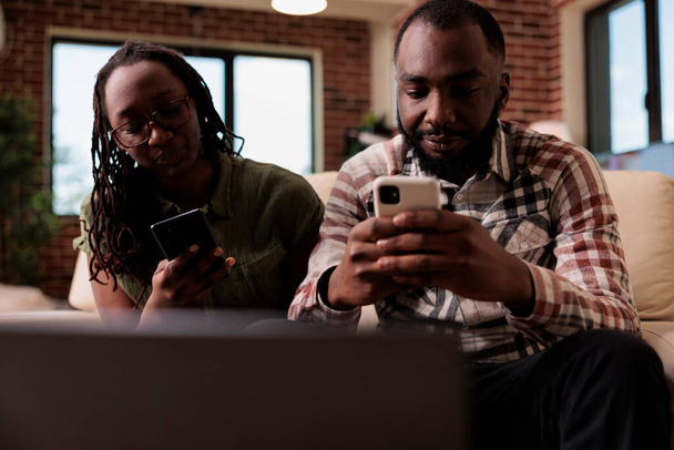 アフリカ系アメリカ人のカップルは、ソファに座っているスマートフォンで過ごす時間がお互いに話をすることができない問題があります。彼氏無視動揺ガールフレンド後に議論でホームリビングルーム. - 写真・画像