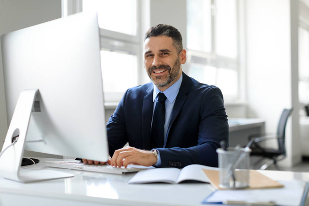 Счастливый бизнесмен средних лет в костюме сидит на рабочем месте в офисе и улыбается в камеру, успешный мужчина-предприниматель, работающий с современным компьютером, свободное пространство - Фото, изображение