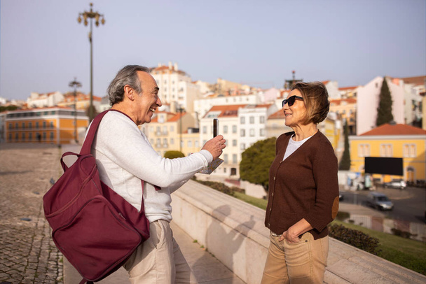 Dovolená. Šťastný starší manžel fotografování své ženy přes Smartphone stojící s batohem venku. Evropský pár seniorů se těší na životní cestování do důchodu a baví se v Lisabonu - Fotografie, Obrázek