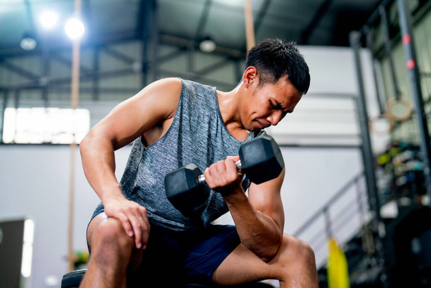Asian sport man sit on chair and hold and lift dumbbell up with one hand and look to arm muscle in fitness gym and look concentrate to training. - Photo, image