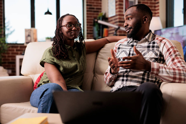 Afro-Amerikaans echtpaar praten met elkaar terwijl ze op de bank zitten in de woonkamer. Vriendin kijkt ontevreden op vriendje uit te leggen en de hand gebaren in de voorkant van de tafel met laptop. - Foto, afbeelding